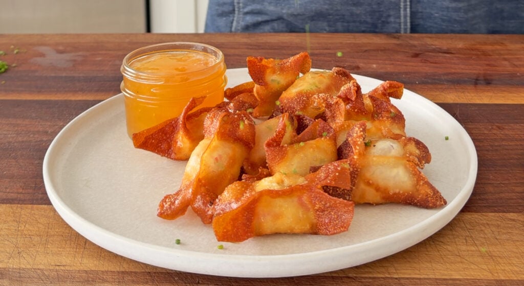 Crispy crab rangoon garnished with sliced chives and served with a bowl of duck sauce for dipping